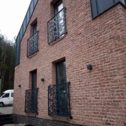 Elewacja budynku z czerwonej cegły z nowoczesnymi, metalowymi balustradami balkonowymi o geometrycznym wzorze, oświetlona przez małe, kwadratowe lampy ścienne, z widocznym fragmentem lasu w tle.