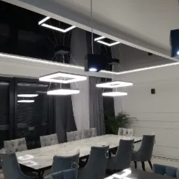 Nowoczesna jadalnia z długim stołem z marmurowym blatem, szarymi krzesłami z weluru i designerskimi lampami wiszącymi o kwadratowym kształcie, czarny sufit podwieszany z listwami LED.