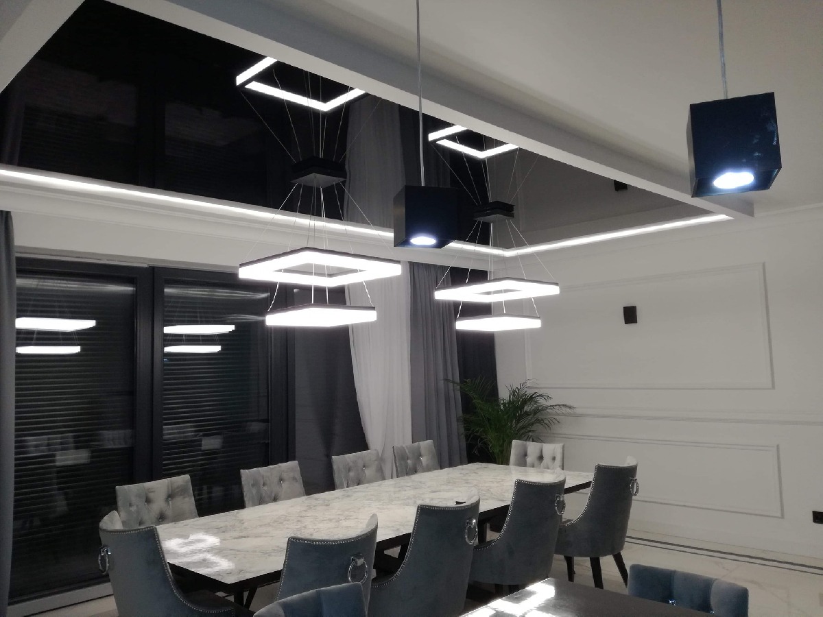 Nowoczesna jadalnia z długim stołem z marmurowym blatem, szarymi krzesłami z weluru i designerskimi lampami wiszącymi o kwadratowym kształcie, czarny sufit podwieszany z listwami LED.