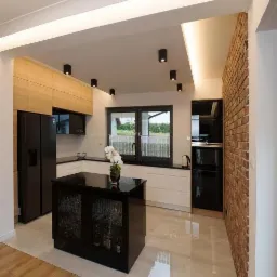 Nowoczesna kuchnia z czarną wyspą, szklanymi frontami i oświetleniem punktowym, uzupełniona o ścianę z cegły dekoracyjnej.