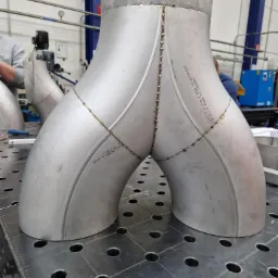 Połączenie spawane dwóch rur ze stali nierdzewnej w kształcie litery Y, widoczne ślady spawania punktowego, całość umieszczona na perforowanym stole spawalniczym, w tle widoczny sprzęt spawalniczy.