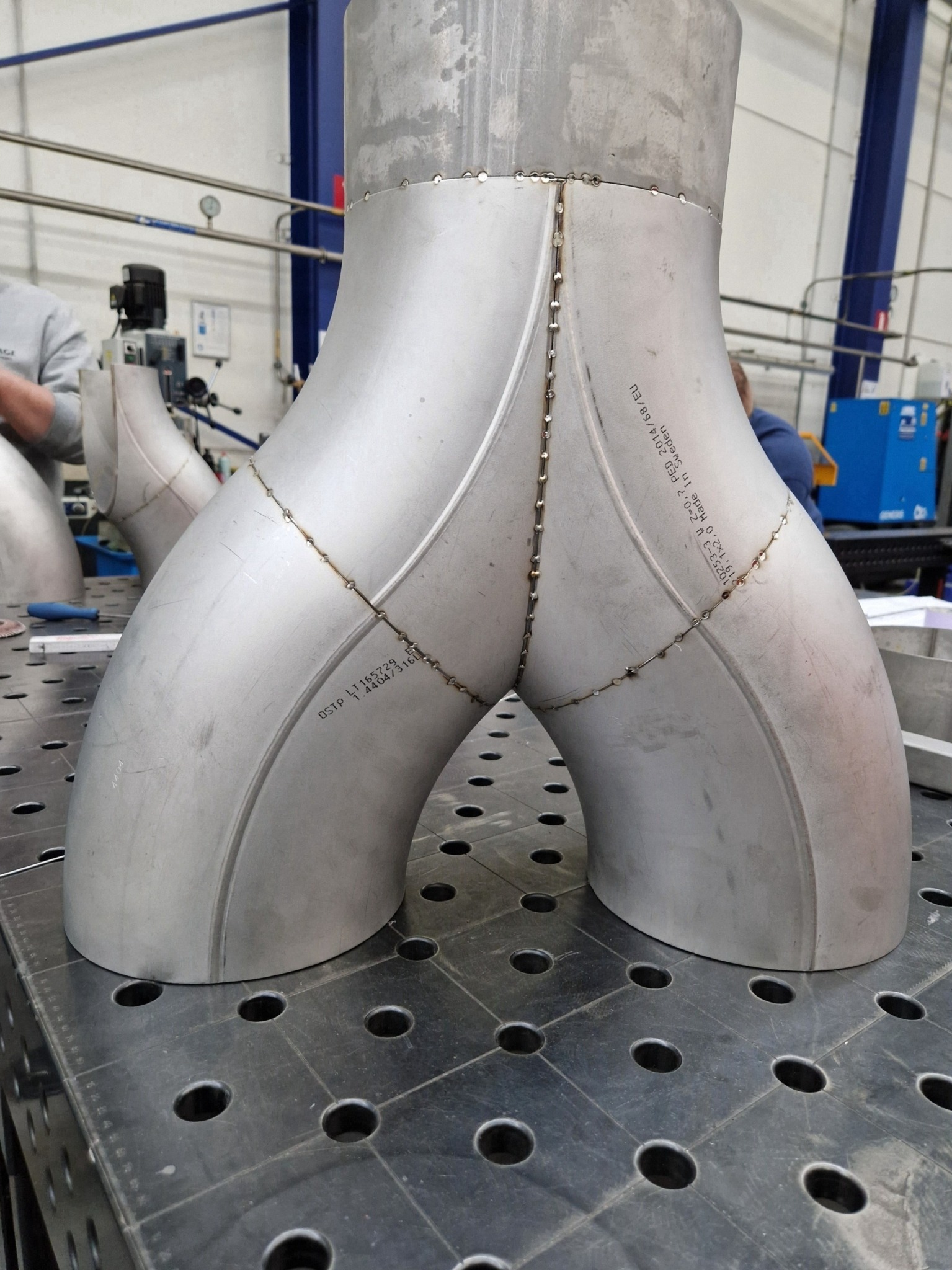 Połączenie spawane dwóch rur ze stali nierdzewnej w kształcie litery Y, widoczne ślady spawania punktowego, całość umieszczona na perforowanym stole spawalniczym, w tle widoczny sprzęt spawalniczy.