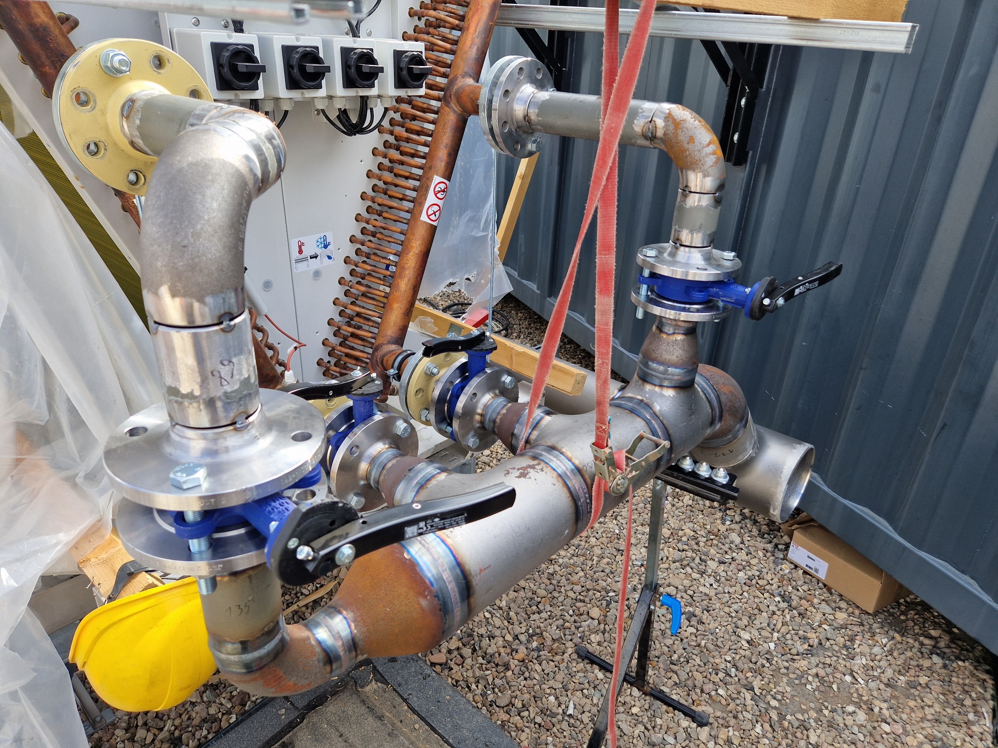 Kompleksowy układ rur stalowych z zaworami i kołnierzami, połączonych spawami, widoczny żółty kask na ziemi, sugerujący trwające prace instalacyjne.