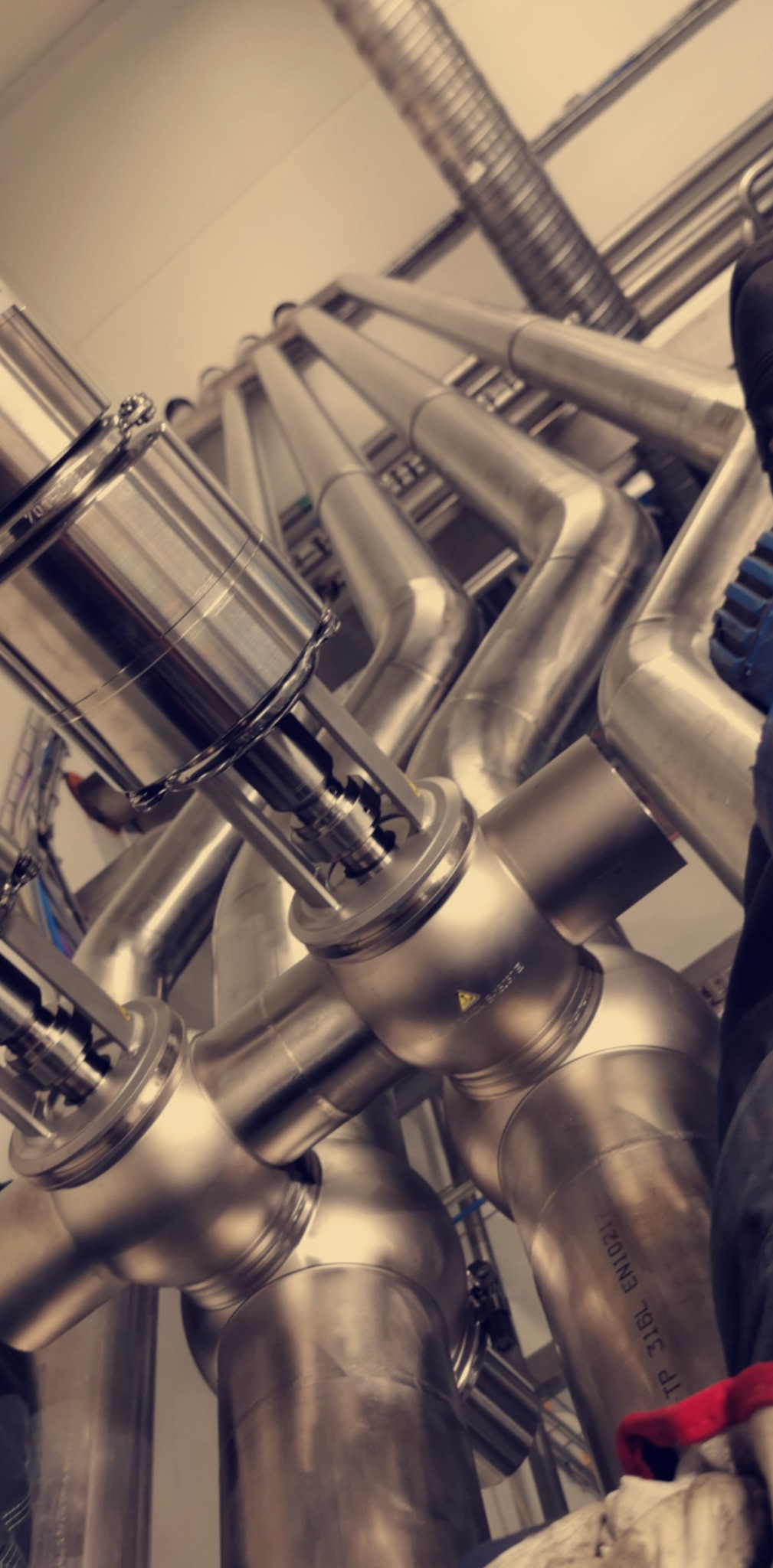 Złożony system rur ze stali nierdzewnej z zaworami, widoczne oznaczenia i elementy mocujące, ujęcie z dołu, fragment roboczego ubrania w kadrze.