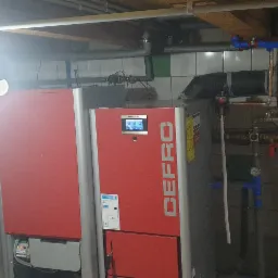 Czerwono-szary piec grzewczy Defro w piwnicy, z widocznymi rurami instalacji grzewczej i oświetleniem roboczym