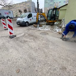 TomWer -Instalacje hydrauliczne, gazowe, wodne i kanalizacyjne, Usługi minikoparką Tomasz Kostuń - Pracownik w niebieskiej kurtce przecina chodnik szlifierką kątową w pobliżu studzienki, na tle zaparkowany biały bus i żółta mini koparka, widoczne barierki drogowe.