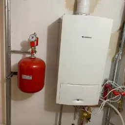 Biały piec gazowy Junkers z podłączonym czerwonym naczyniem przeponowym i instalacją hydrauliczną na tle ściany z widocznymi śladami po remoncie.