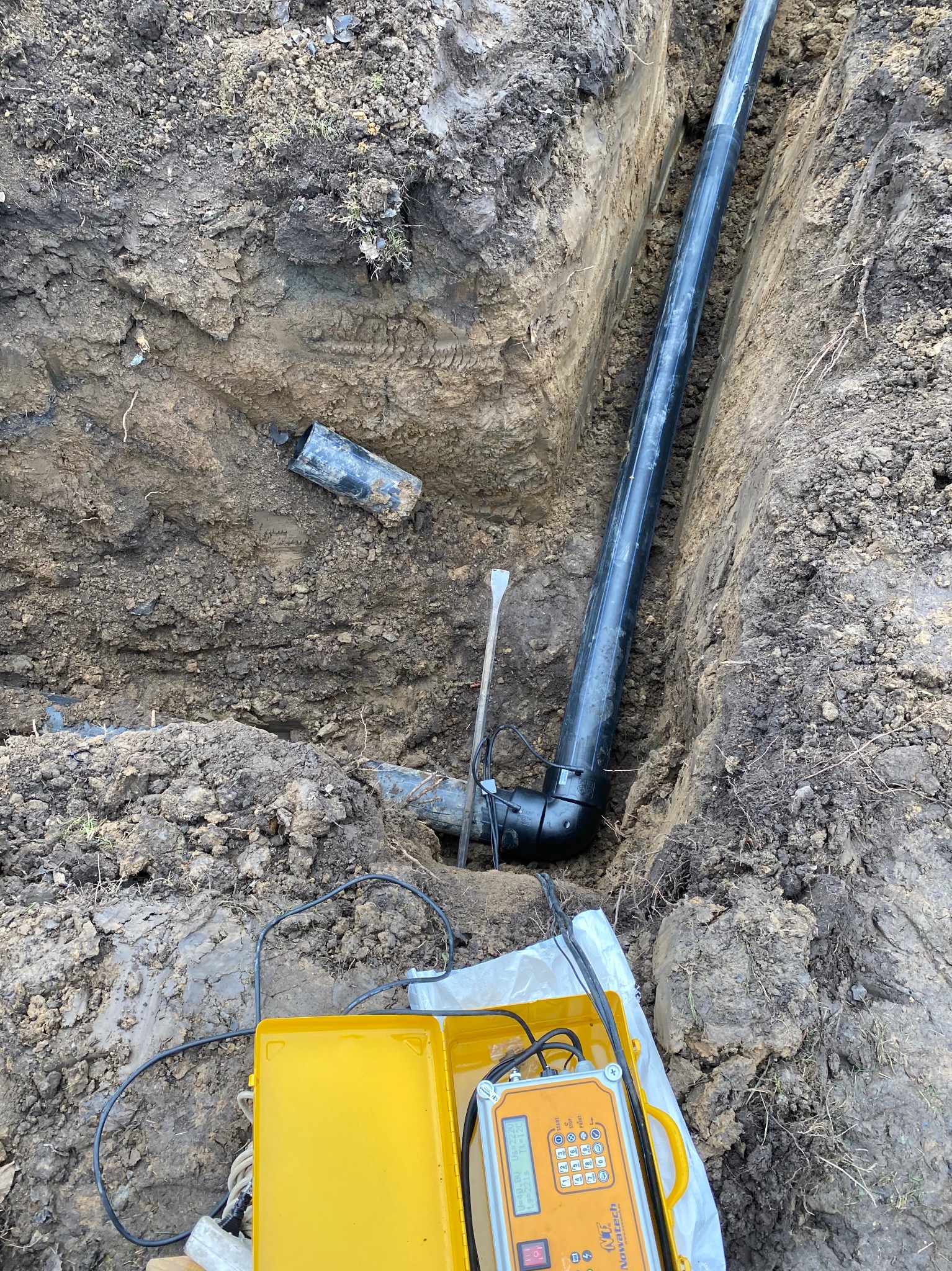 Rów z ułożoną czarną rurą kanalizacyjną, widoczny fragment starej rury i żółte urządzenie pomiarowe Nowatech z ekranem LCD, ziemia gliniasta.