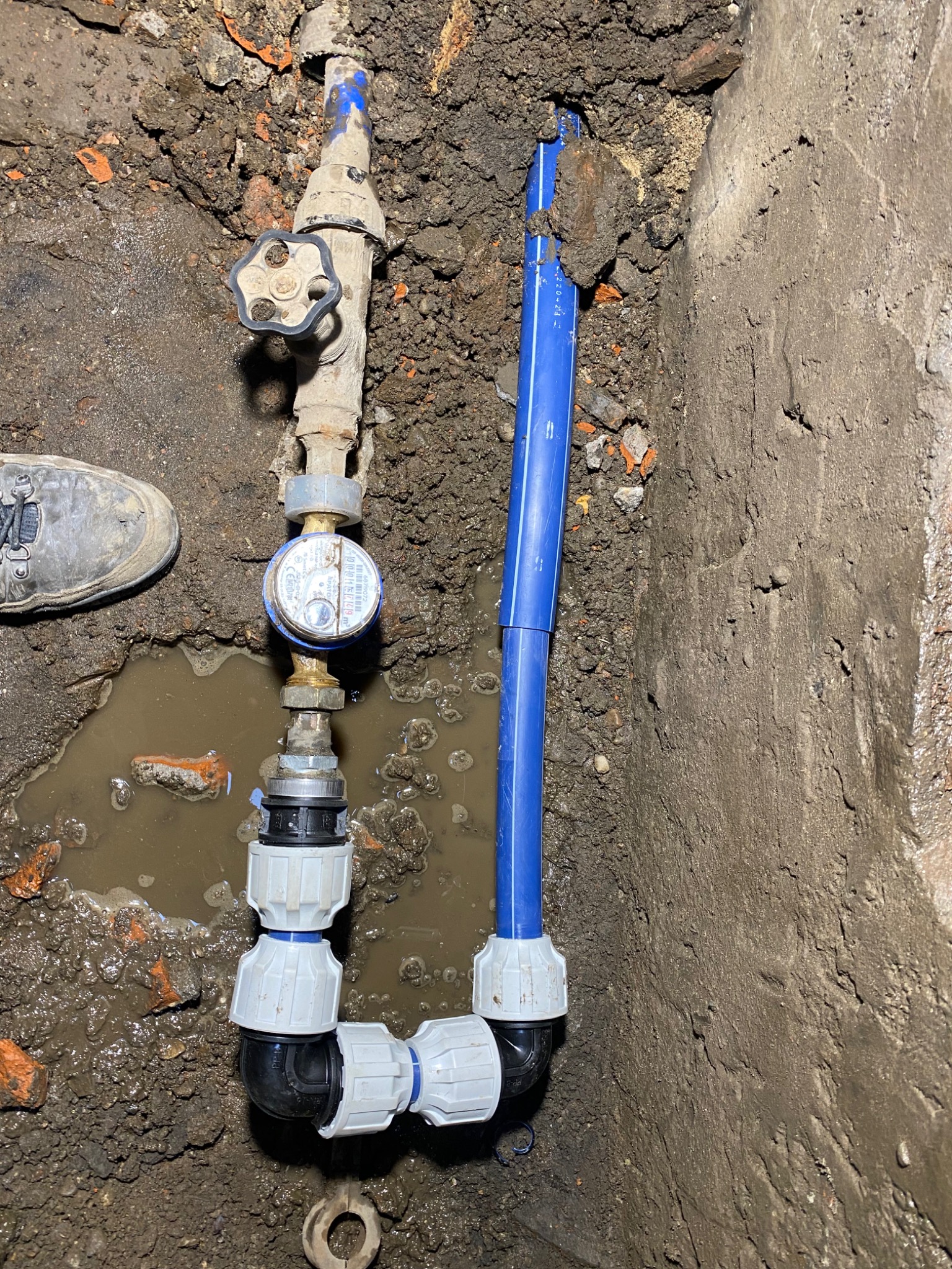 Instalacja wodociągowa z licznikiem w wykopie, widoczne złącza, zawór i fragment buta pracownika na błotnistym gruncie.