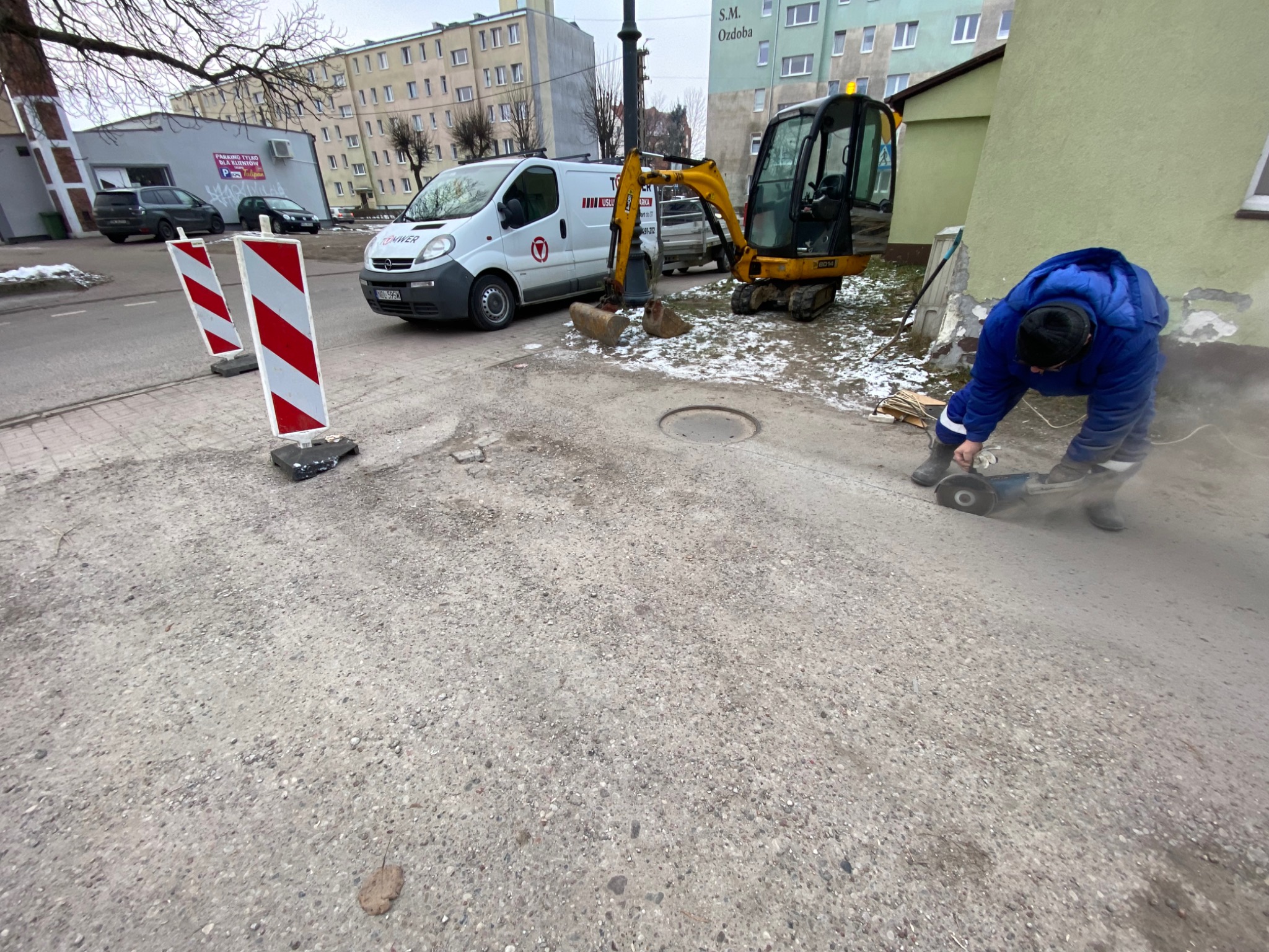 Pracownik w niebieskiej kurtce przecina chodnik szlifierką kątową w pobliżu studzienki, na tle zaparkowany biały bus i żółta mini koparka, widoczne barierki drogowe.