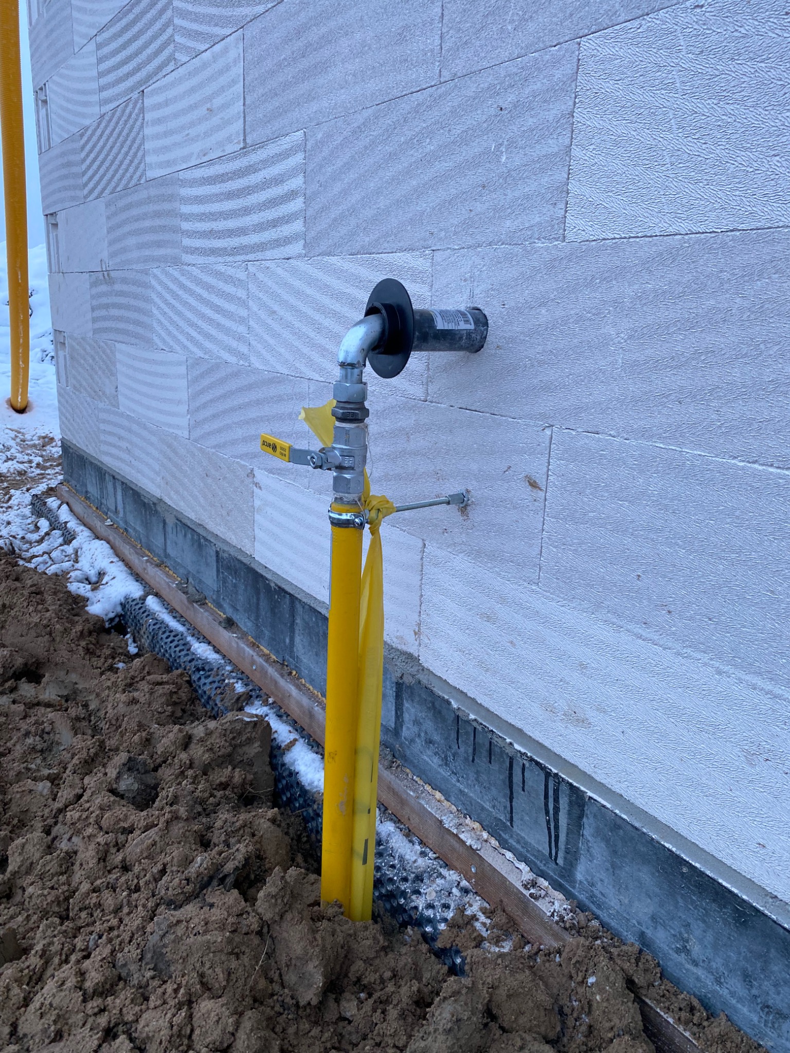 Żółta rura gazowa wystająca z ziemi przy ścianie budynku z betonu komórkowego, widoczny zawór odcinający i czarny kołnierz na ścianie, fragment ośnieżonej ziemi.