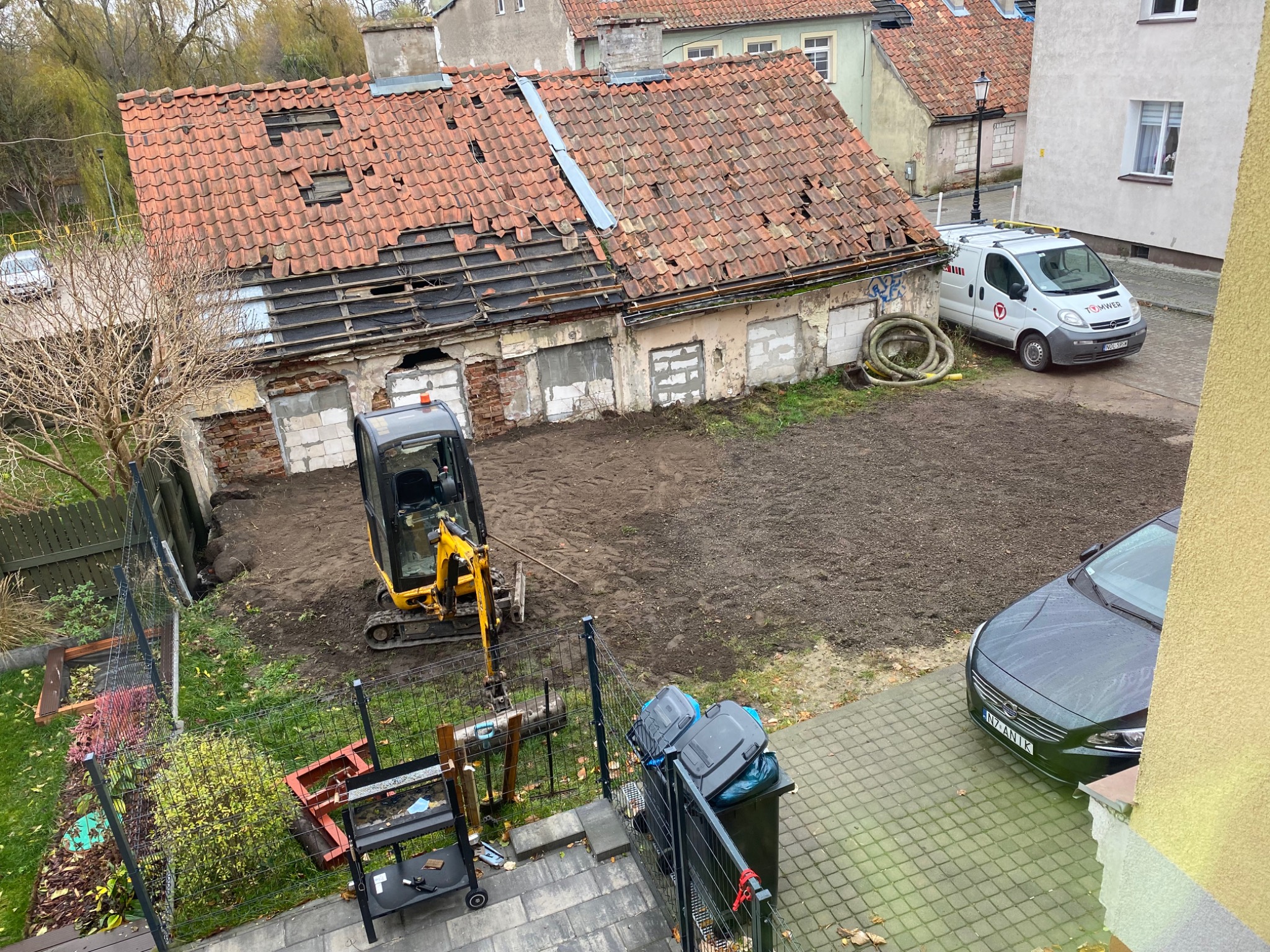 Żółta mini koparka na działce z przygotowanym terenem pod budowę, w tle zrujnowany budynek z dziurawym dachem z czerwonej dachówki i zaparkowany biały samochód dostawczy.