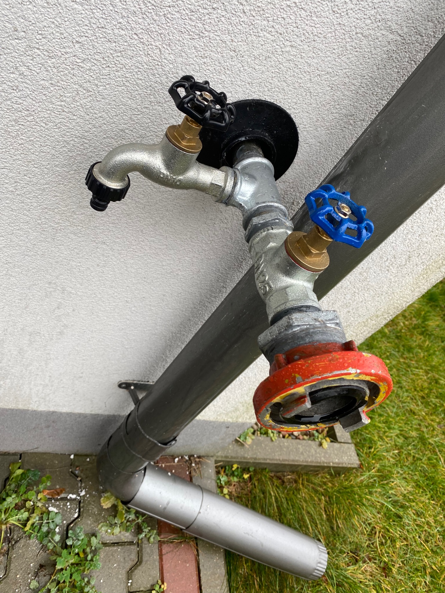 Zewnętrzny zawór wodny z czarnym i niebieskim pokrętłem, zamontowany na ścianie budynku obok rury spustowej, widoczny fragment trawnika.
