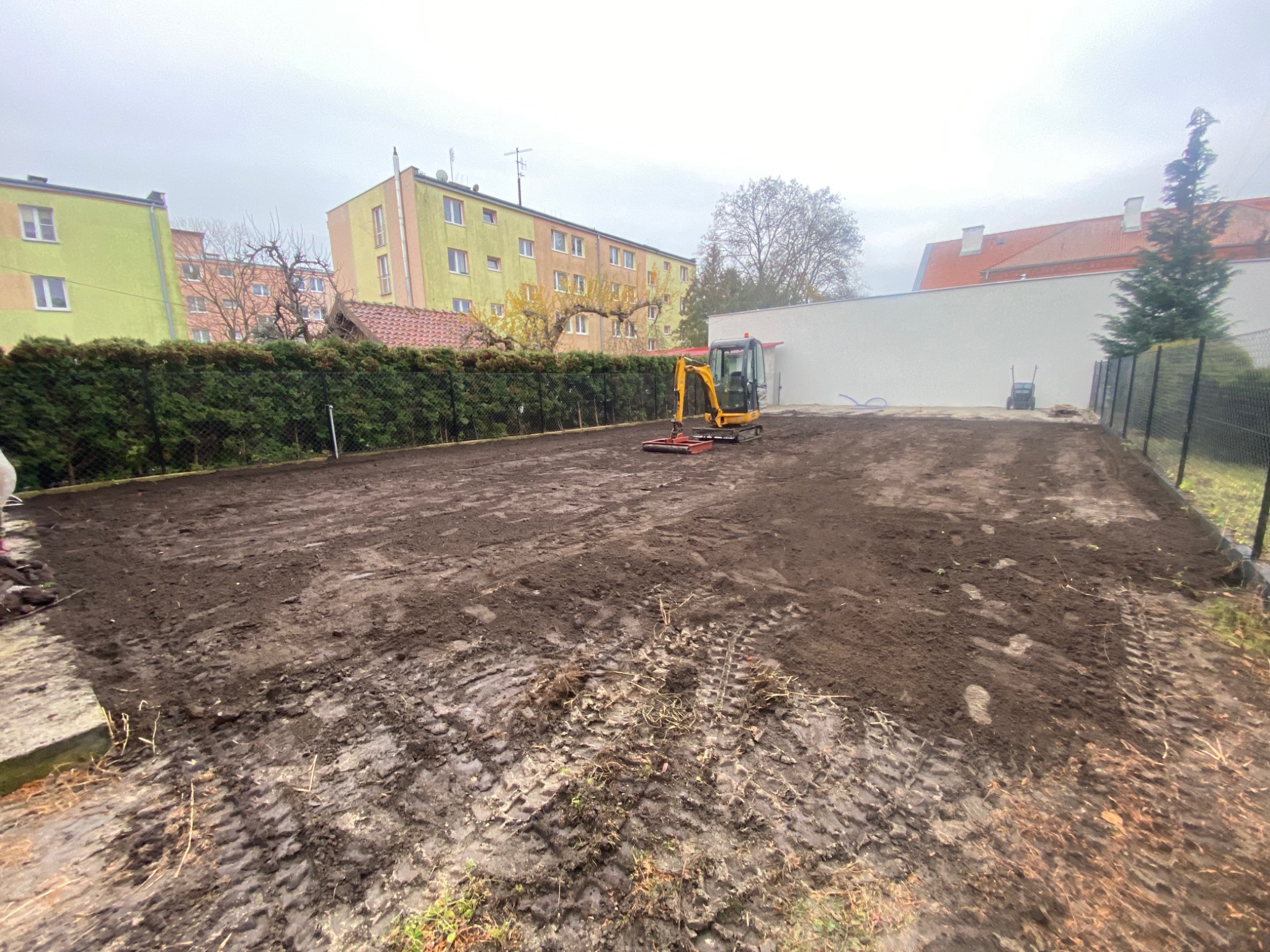 Żółta mini koparka na wyrównanym, błotnistym terenie przygotowanym pod budowę, w tle budynki mieszkalne i zielony żywopłot.