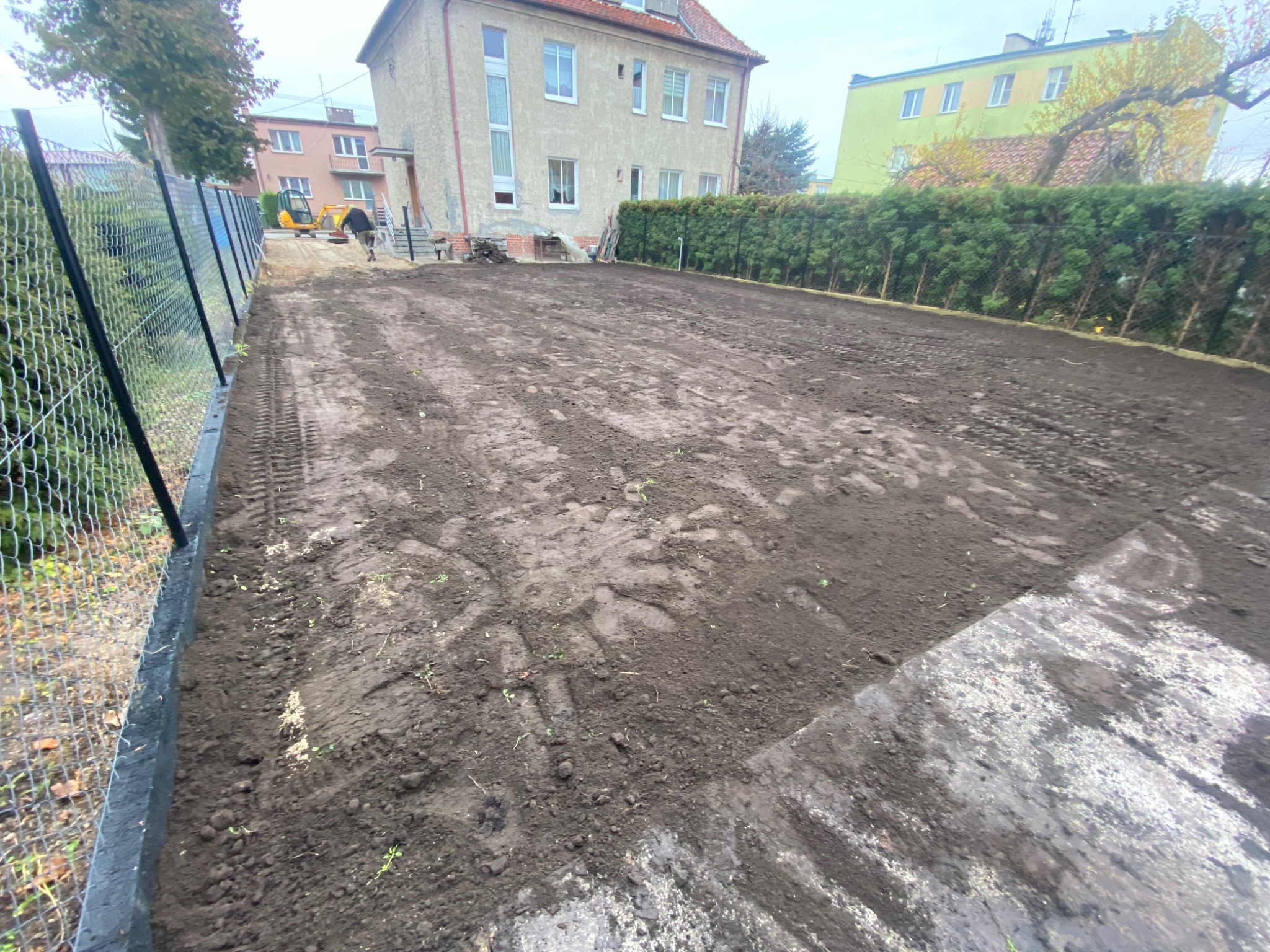 Przygotowany teren pod budowę z widocznymi śladami opon i pracującą koparką w tle, ogrodzony siatką.
