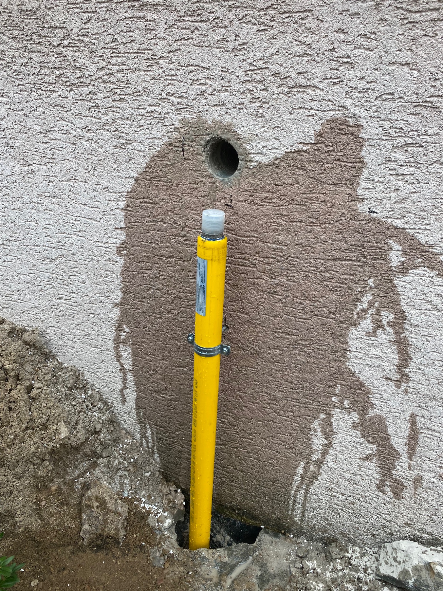 Żółta rura wystająca z ziemi przy ścianie budynku z otworem, widoczne ślady po cieczy na elewacji.