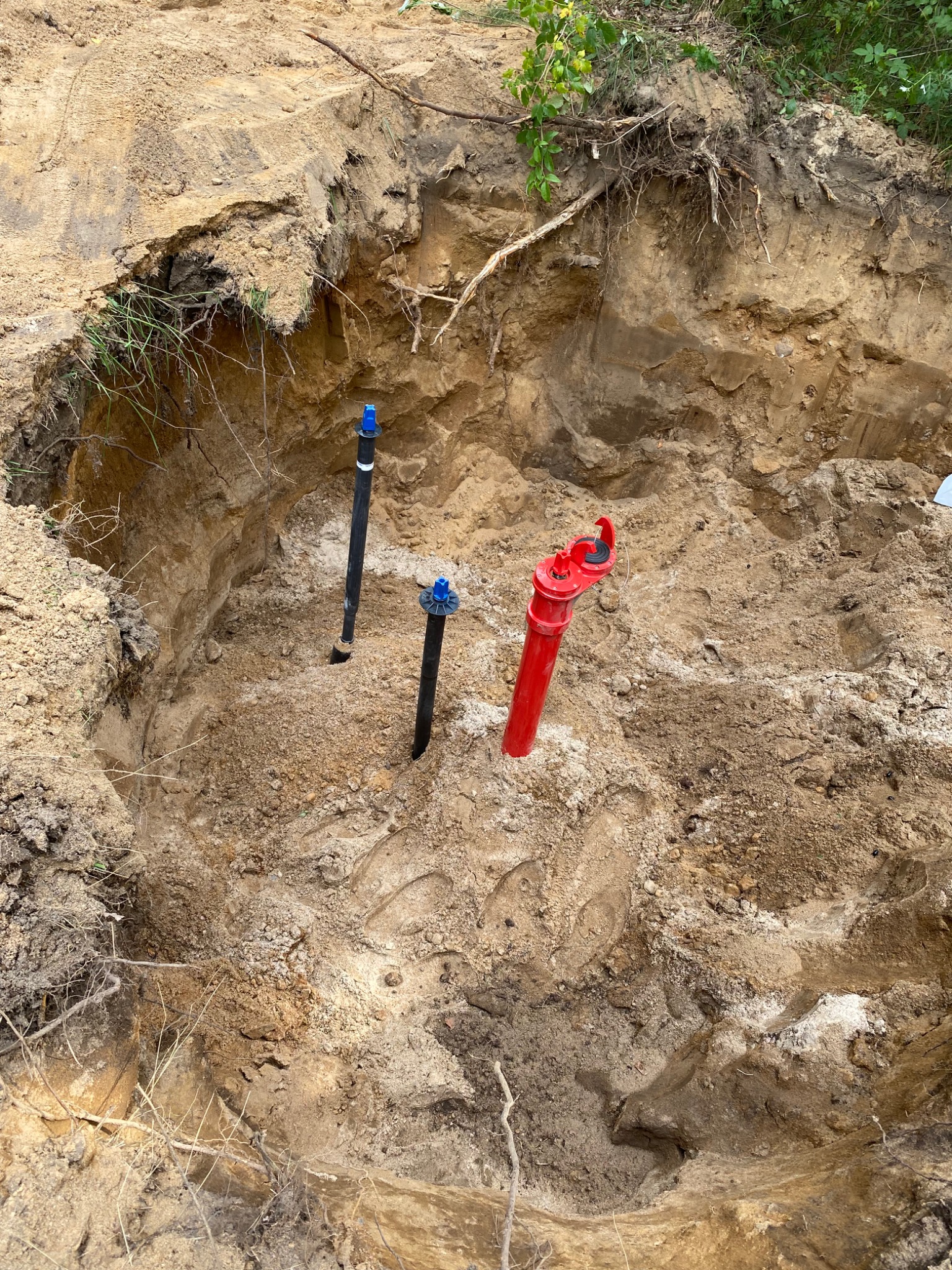 Głęboki wykop w ziemi z wystającymi rurami: czarne rury z niebieskimi końcówkami i czerwona rura z zaworem, widoczne ślady stóp na dnie wykopu, korzenie drzew na krawędzi wykopu.