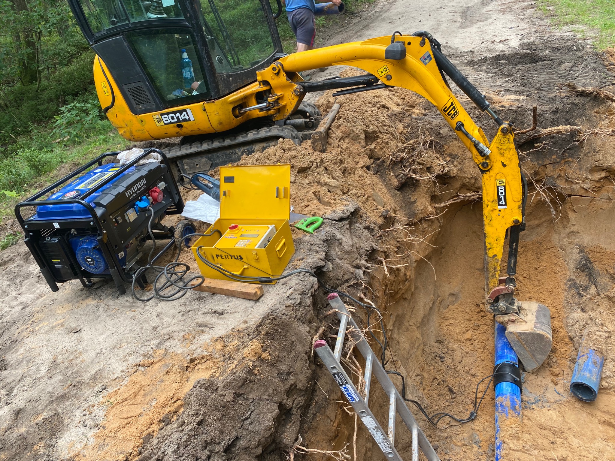 Żółta koparka JCB z niebieskim agregatem prądotwórczym Hyundai obok wykopu z widoczną rurą wodociągową i żółtą skrzynką pomiarową Pertus oraz drabina Krause w wykopie.