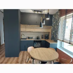 Nowoczesna kuchnia w odcieniach szarości i drewna z okrągłym stołem i krzesłami. Na ścianie widoczna tapeta w roślinny wzór, a nad blatem wiszą trzy czarne lampy ze złotymi elementami.