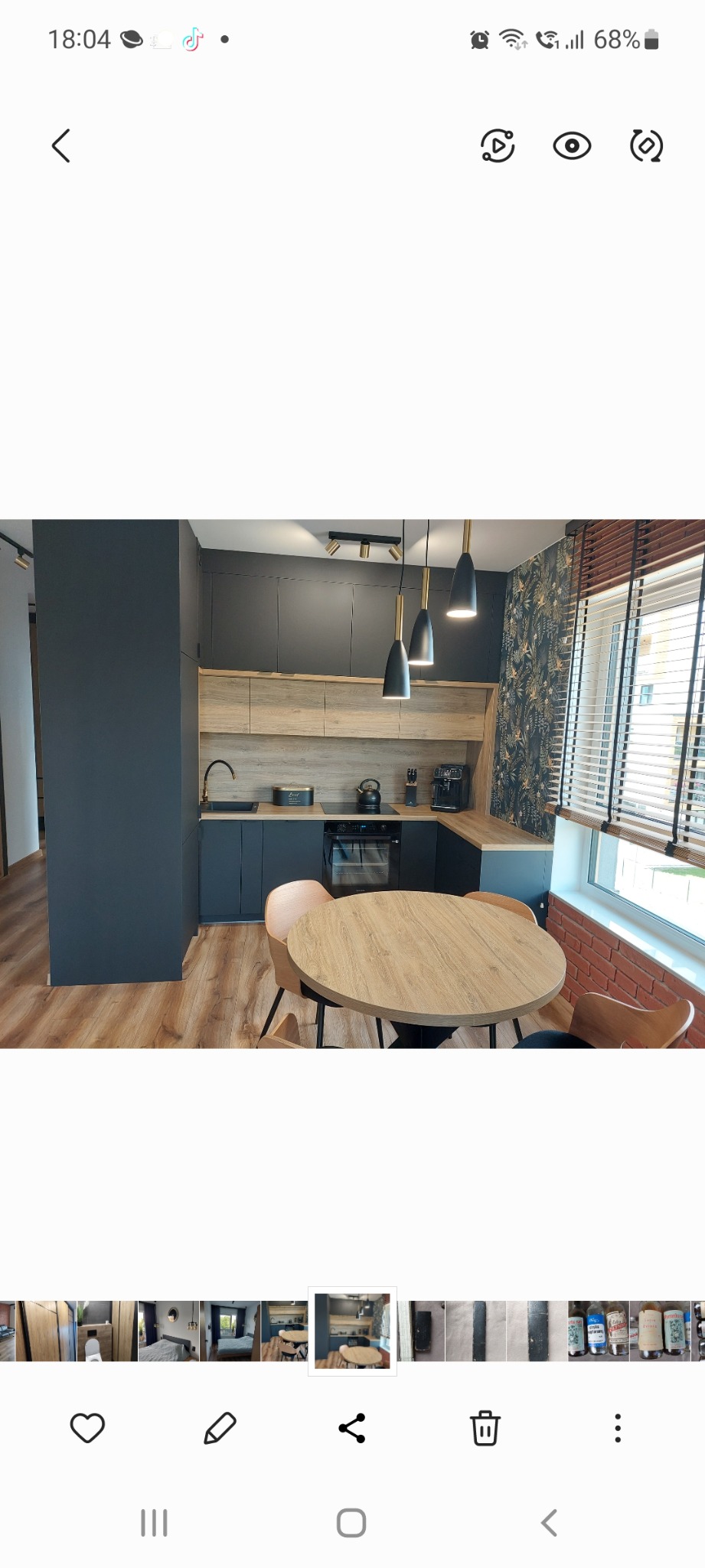 Nowoczesna kuchnia w odcieniach szarości i drewna z okrągłym stołem i krzesłami. Na ścianie widoczna tapeta w roślinny wzór, a nad blatem wiszą trzy czarne lampy ze złotymi elementami.