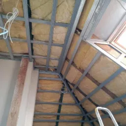 Montaż metalowej konstrukcji pod płytę kartonowo-gipsową na poddaszu z widoczną izolacją z wełny mineralnej, częściowo oświetlonym oknem dachowym i fragmentem drabiny.