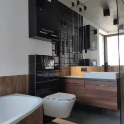 Nowoczesna łazienka z czarnymi, błyszczącymi płytkami na ścianie, drewnianą zabudową wanny i szafki pod umywalką, wiszącą toaletą oraz minimalistycznym oświetleniem punktowym.