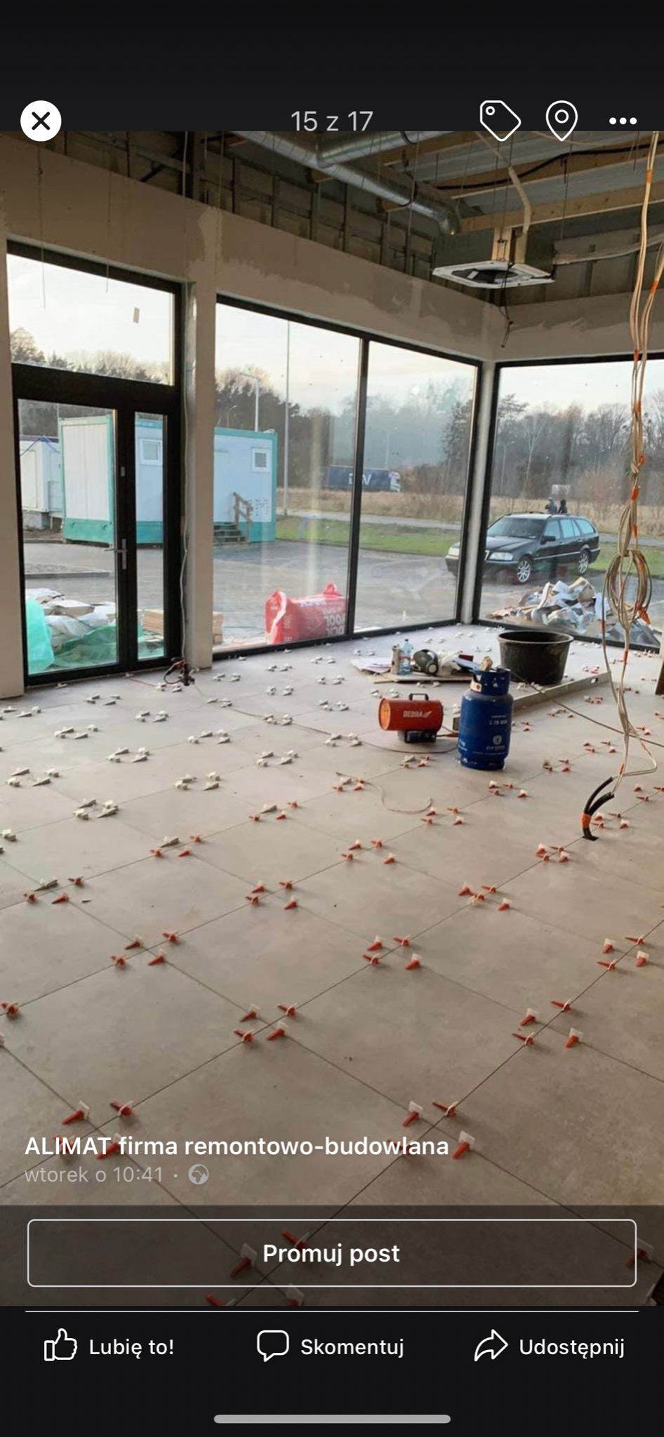 Układanie jasnych płytek podłogowych w dużym pomieszczeniu, widoczne krzyżyki dystansowe, nagrzewnica, butla gazowa, wiadro i narzędzia, przez okna widać budynek kontenerowy i samochód.