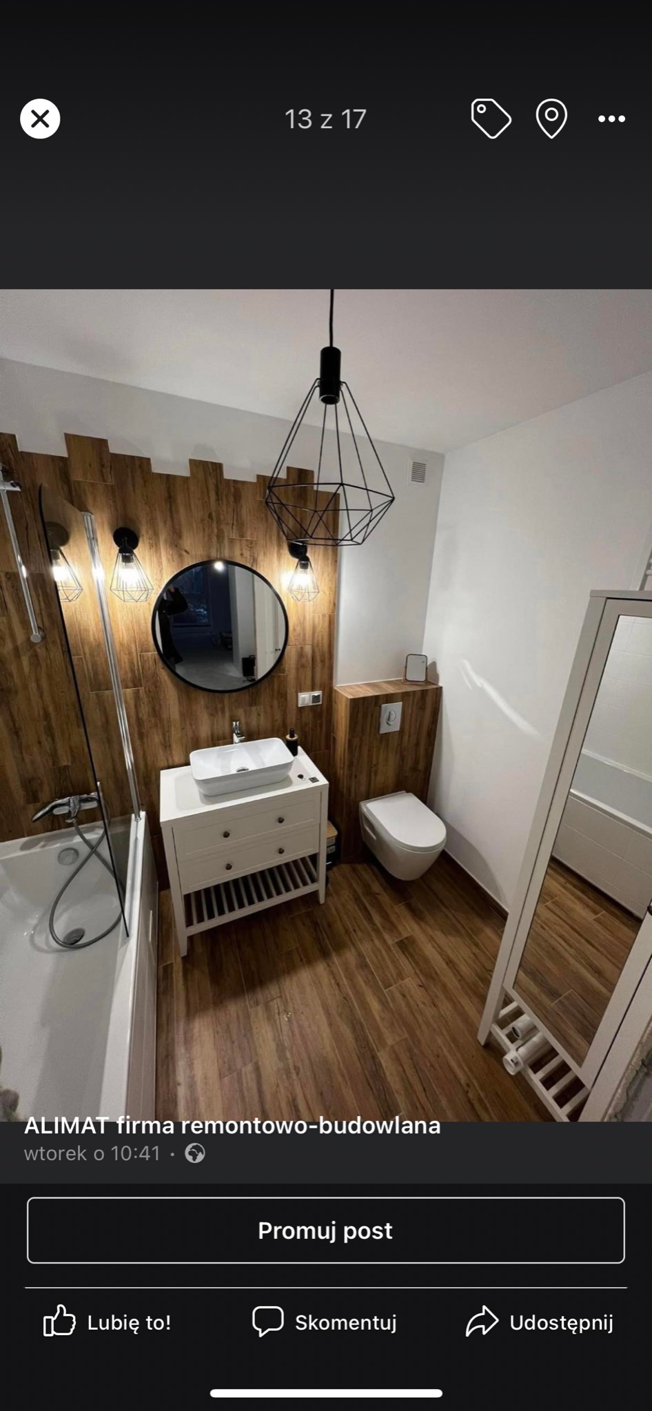 Wykończona łazienka z drewnopodobnymi płytkami na ścianie i podłodze, białą umywalką na szafce z szufladami, okrągłym lustrem w czarnej ramie i geometryczną lampą wiszącą.