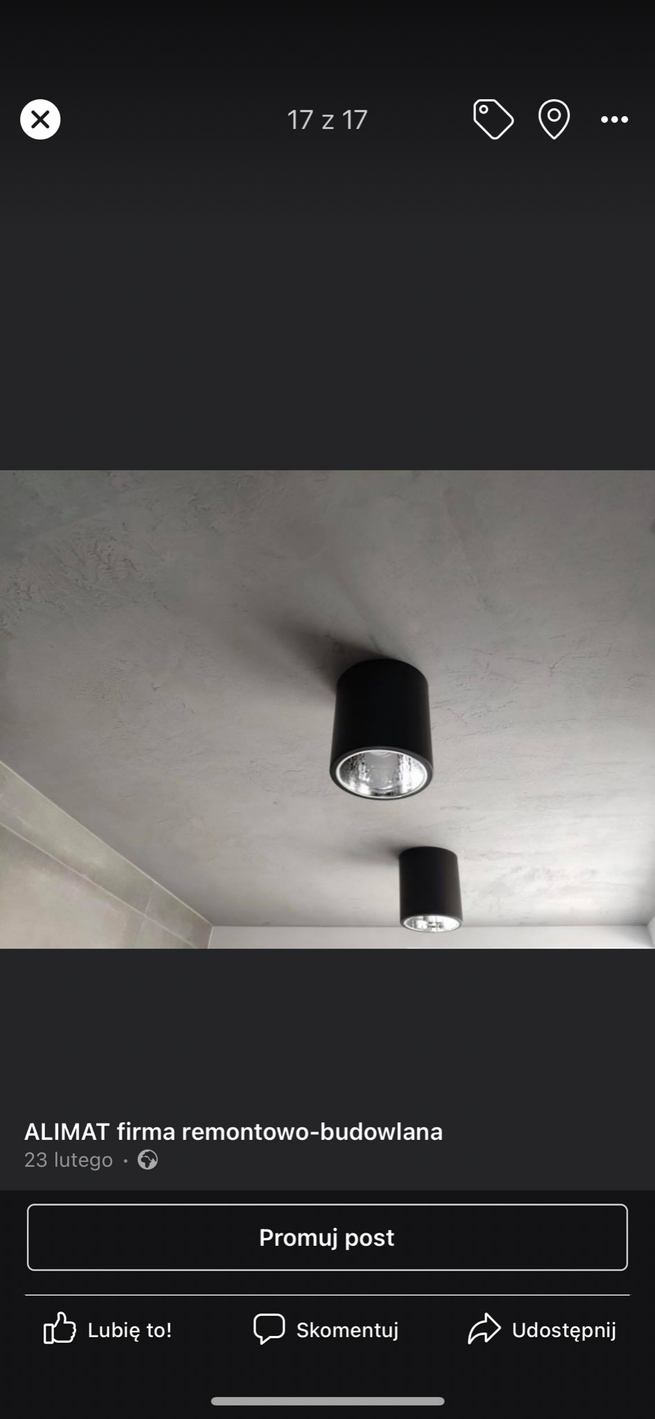 Dwa czarne, cylindryczne reflektory sufitowe w łazience z szarym, teksturowanym sufitem i częściowo widoczną ścianą wyłożoną szarymi płytkami.