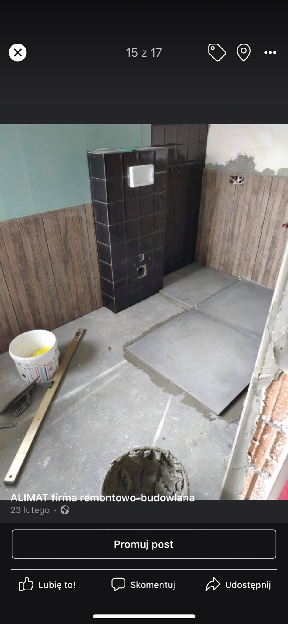 Łazienka w trakcie remontu: czarne płytki na ścianie obok instalacji, drewnopodobne panele ścienne, szare płytki podłogowe w trakcie układania, wiadro z zaprawą i poziomica na podłodze.