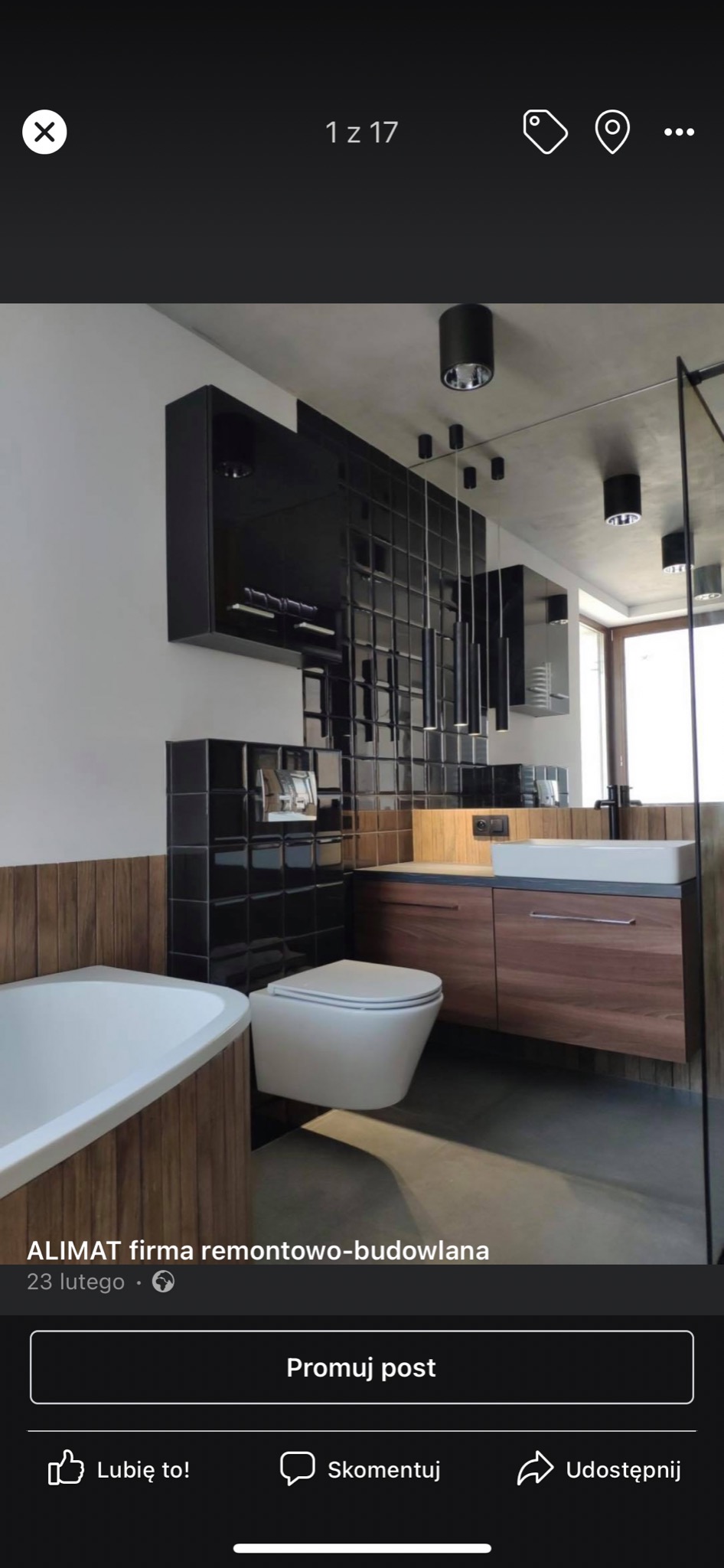 Nowoczesna łazienka z czarnymi, błyszczącymi płytkami, drewnianymi akcentami oraz wiszącą toaletą i umywalką na szafce z drewna orzechowego, oświetlona punktowymi lampami sufitowymi.