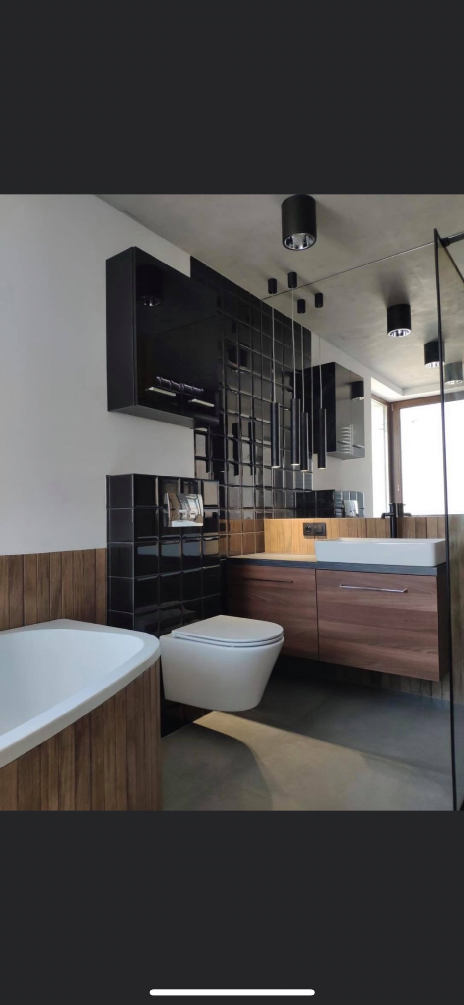 Nowoczesna łazienka z czarnymi, błyszczącymi płytkami na ścianie, drewnianą zabudową wanny i szafki pod umywalką, wiszącą toaletą oraz minimalistycznym oświetleniem punktowym.