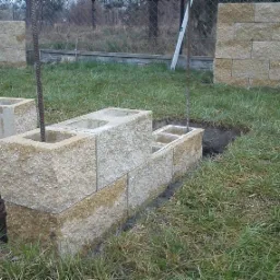 Budowa murku z pustaków betonowych na trawniku, z wystającymi prętami zbrojeniowymi, widoczne wykopy pod fundamenty.