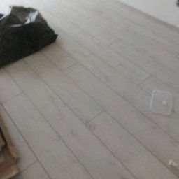 Mar-Bud usługi budowlane - Jasne panele podłogowe ułożone diagonalnie, z widocznym czarnym workiem na śmieci i fragmentem kartonu w rogu, plastikowa osłonka na podłodze.