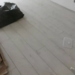 Jasne panele podłogowe ułożone diagonalnie, z widocznym czarnym workiem na śmieci i fragmentem kartonu w rogu, plastikowa osłonka na podłodze.