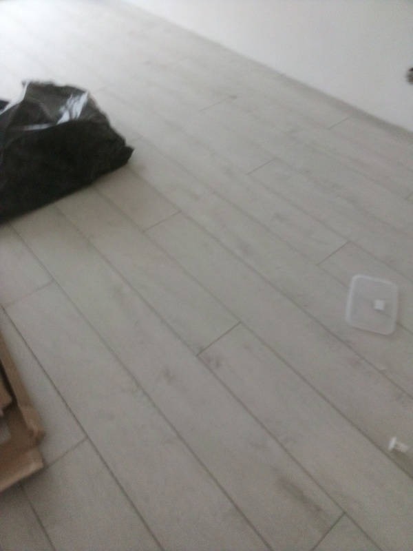 Jasne panele podłogowe ułożone diagonalnie, z widocznym czarnym workiem na śmieci i fragmentem kartonu w rogu, plastikowa osłonka na podłodze.