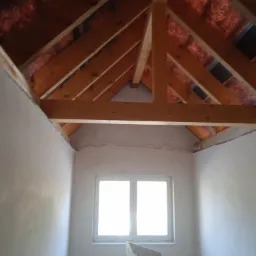 Poddasze w trakcie prac wykończeniowych; widoczne surowe drewniane krokwie, ocieplenie dachu, okno PCV i ściany po wstępnym położeniu gładzi.
