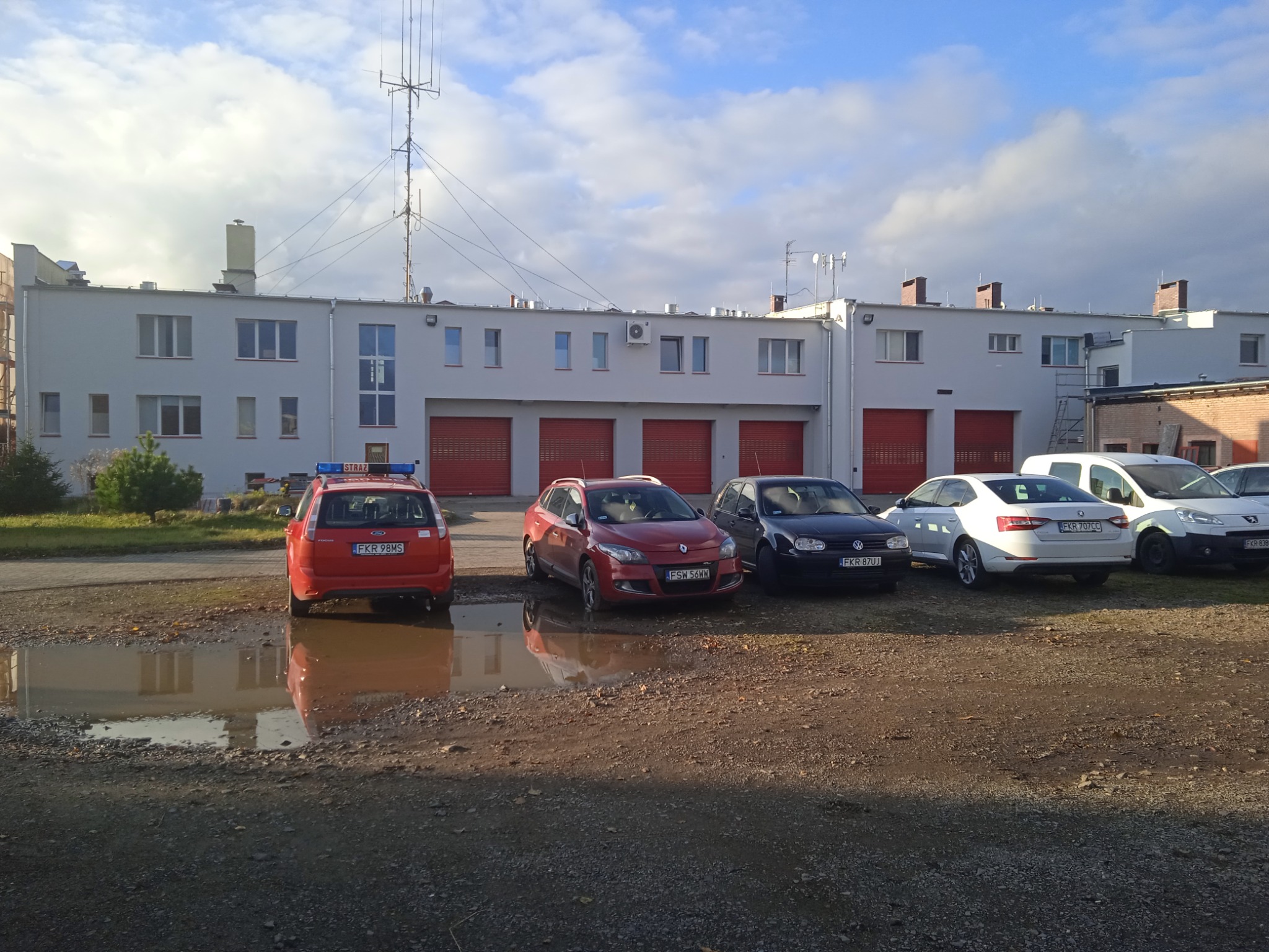 Budynek z czerwonymi bramami garażowymi widziany z zewnątrz, z zaparkowanymi samochodami i kałużą na pierwszym planie, odzwierciedlającą budynek.