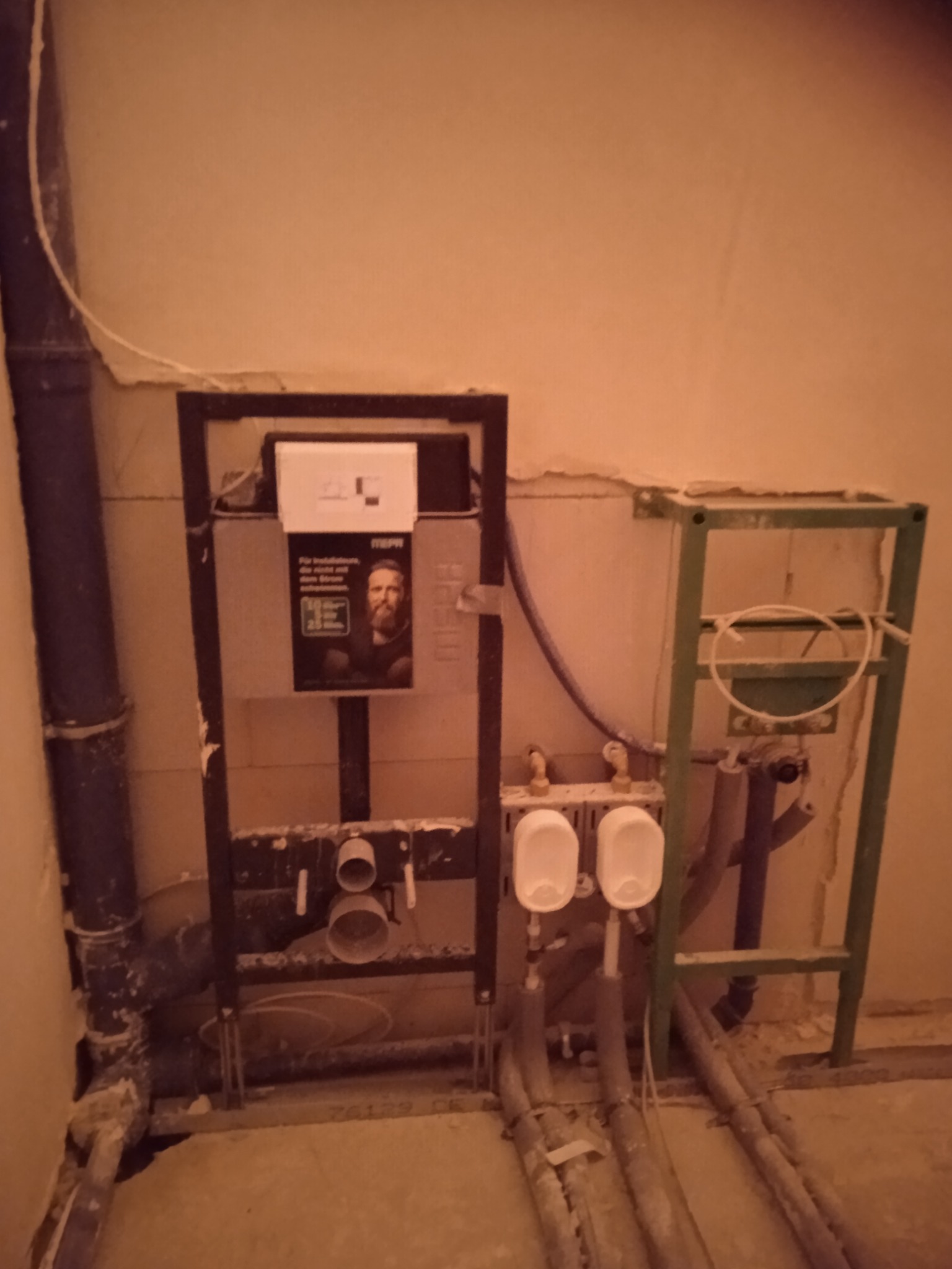 Stelaż podtynkowy do WC z widoczną instalacją hydrauliczną w trakcie montażu, z fragmentem ściany w tle, w surowym stanie.