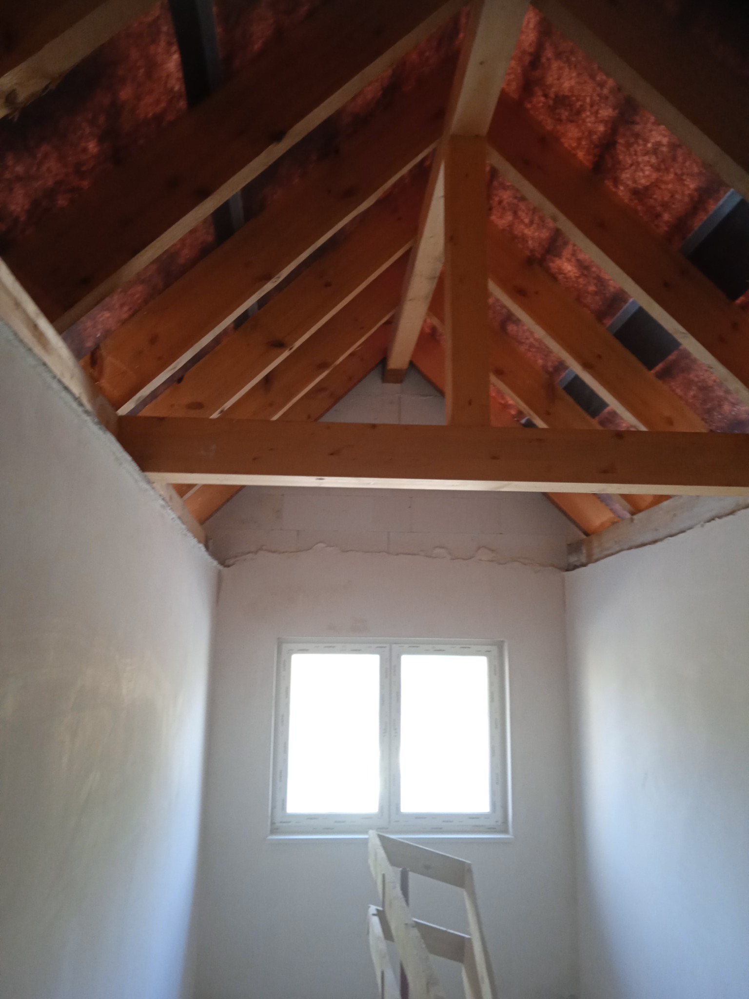 Poddasze w trakcie prac wykończeniowych; widoczne surowe drewniane krokwie, ocieplenie dachu, okno PCV i ściany po wstępnym położeniu gładzi.