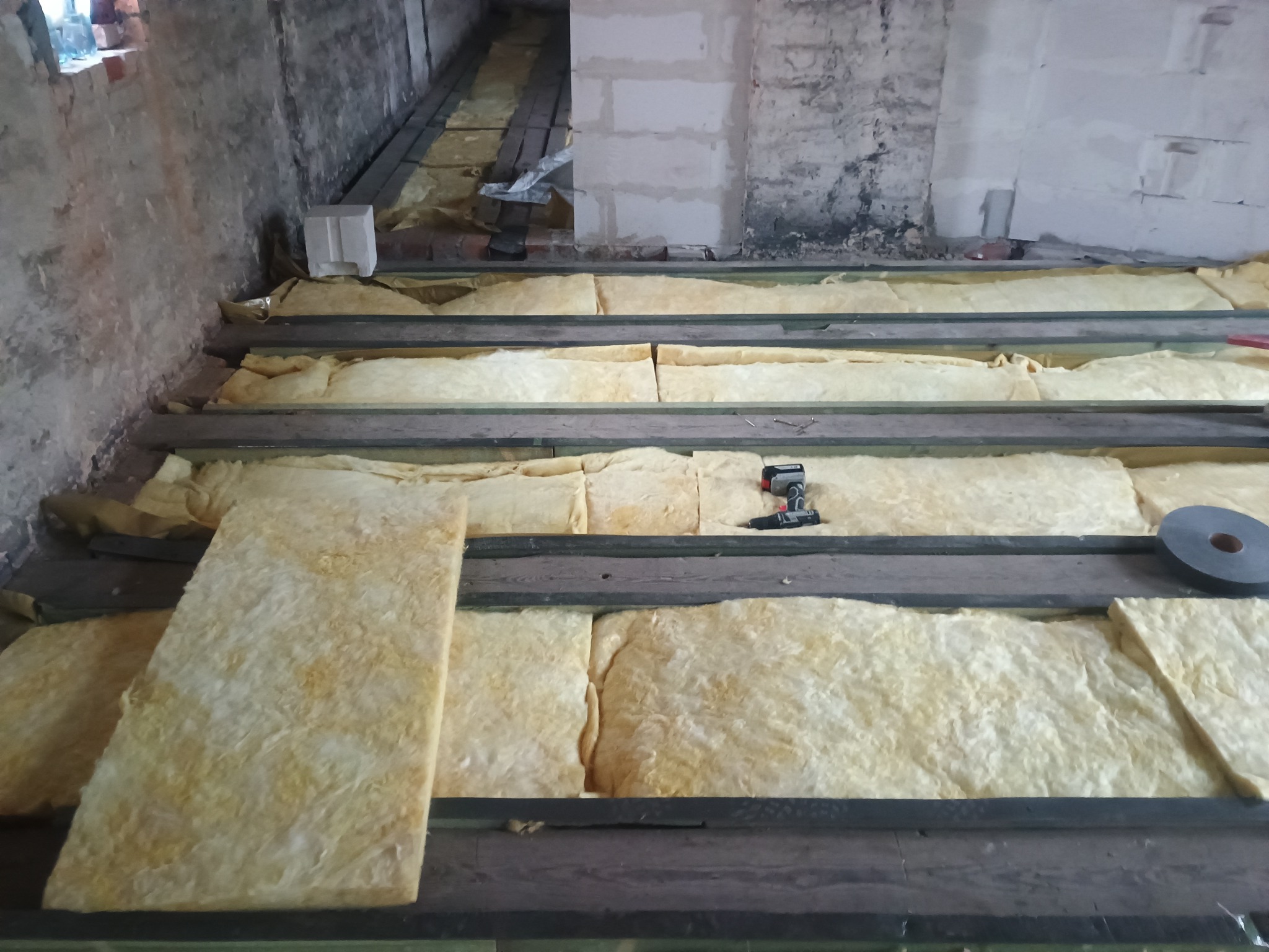 Izolacja podłogi wełną mineralną między legarami, widoczny wkrętak akumulatorowy i taśma izolacyjna. Surowy stan ścian w tle. Prace wykończeniowe w trakcie.