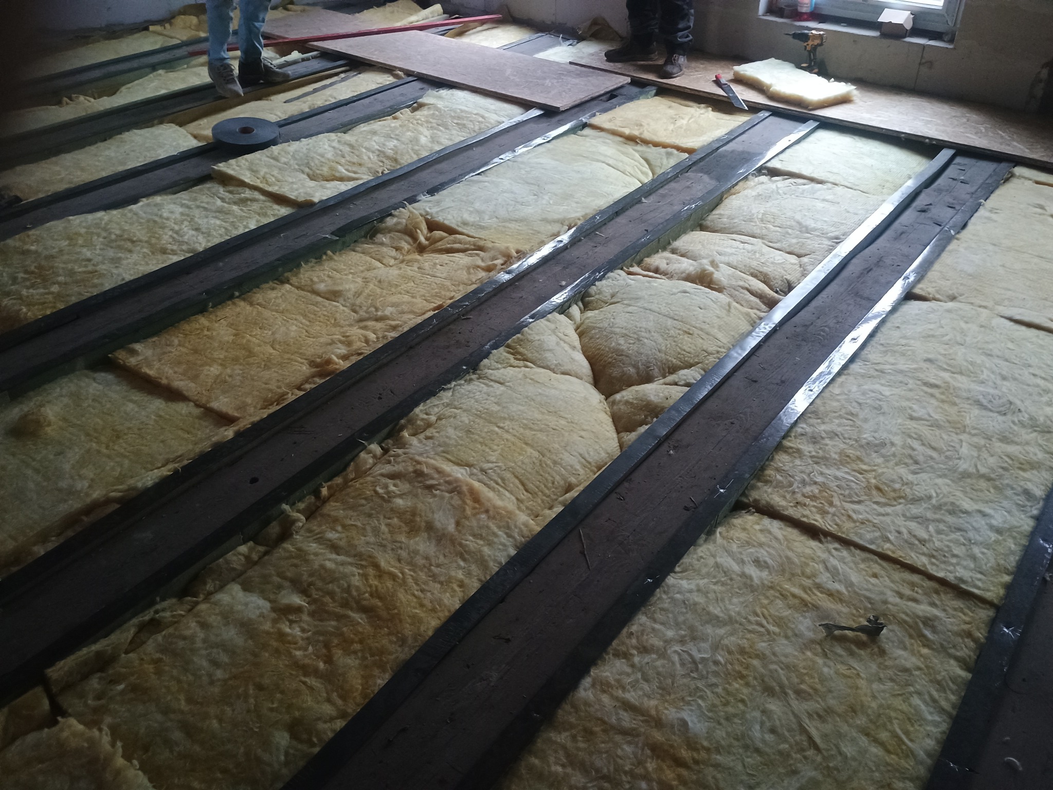 Ocieplenie podłogi wełną mineralną między drewnianymi legarami, widoczne fragmenty podłogi i narzędzia. Etap prac przygotowawczych do położenia podłogi.