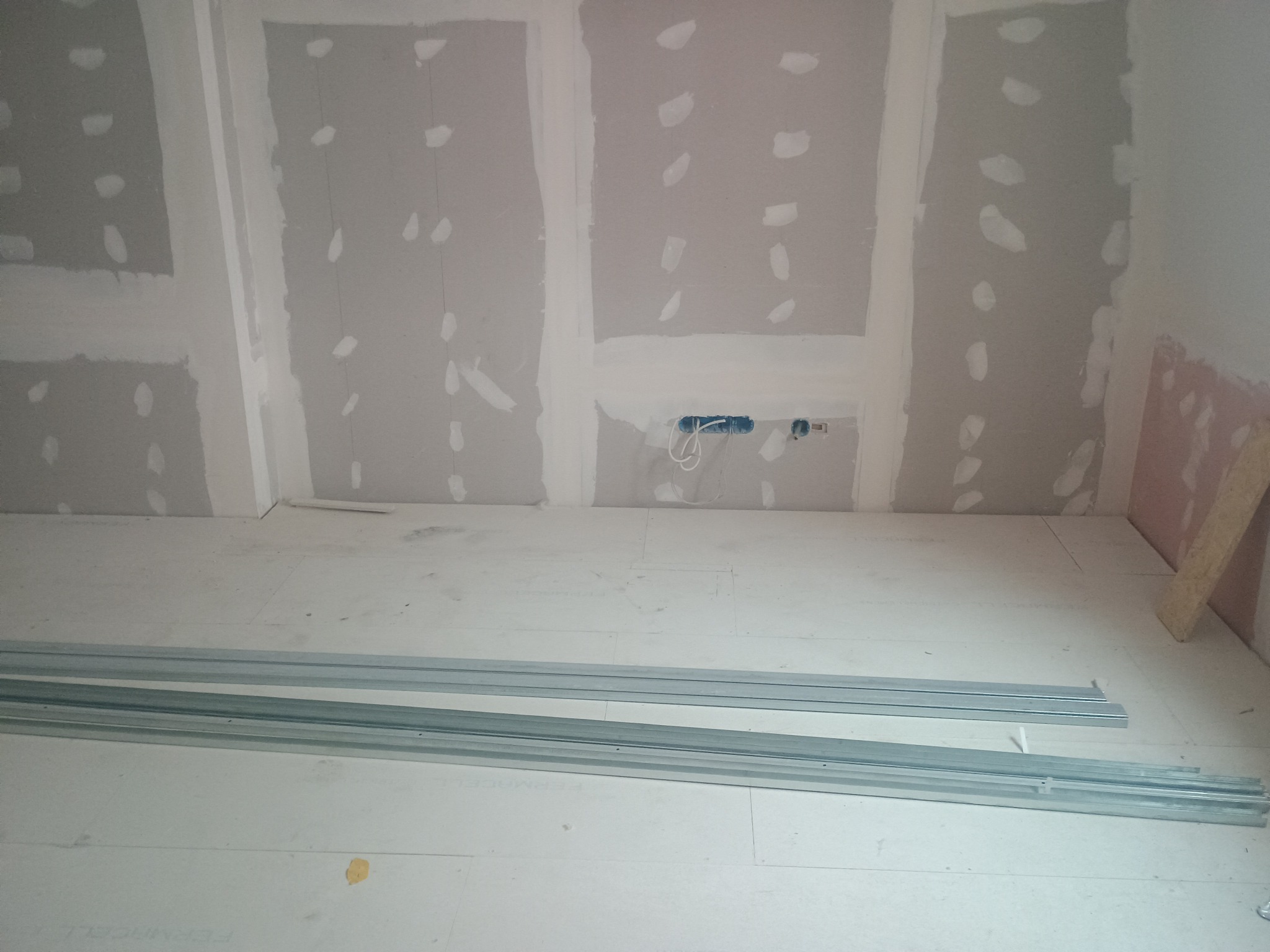 Przygotowanie do montażu płyt gipsowo-kartonowych: widoczne profile na podłodze i ściany z częściowo położoną gładzią, instalacja elektryczna w ścianie.