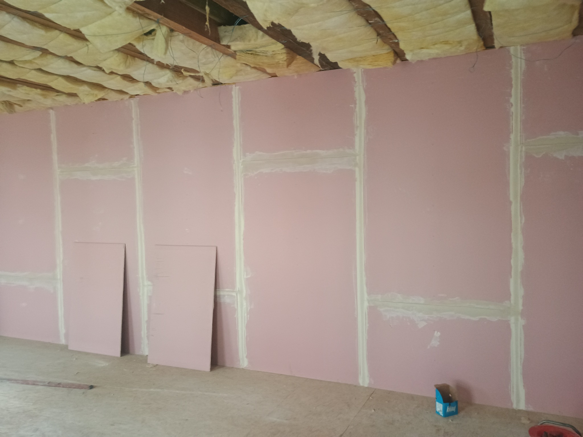 Różowe płyty karton-gips na ścianie, spoiny wypełnione gipsem, izolacja na suficie. Dwie płyty opierają się o ścianę. Widoczny poziomica i pudełko po gipsie.