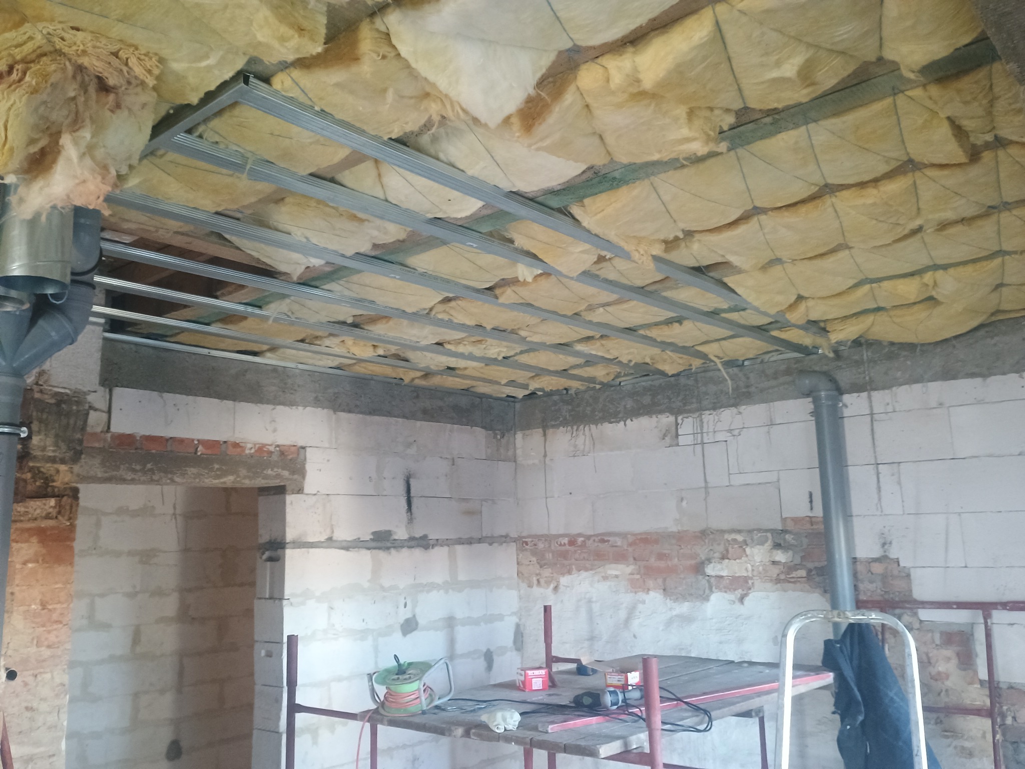 Montaż sufitu podwieszanego z izolacją z wełny mineralnej na metalowej konstrukcji w surowym wnętrzu. Widoczne instalacje i fragment ściany z pustaków.