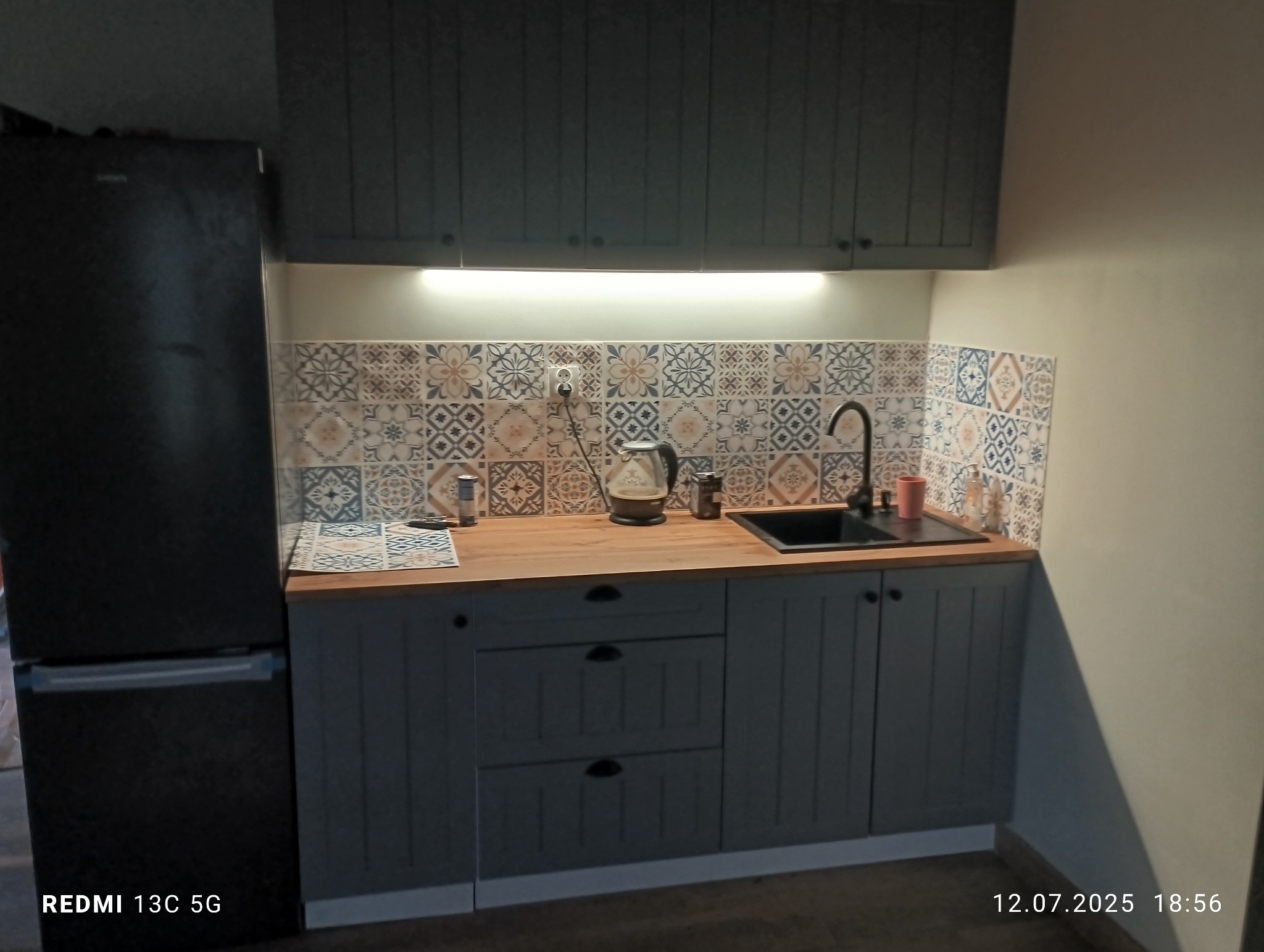 Szara zabudowa kuchenna z drewnianym blatem i wzorzystymi płytkami nad blatem. Widoczny czajnik elektryczny i zlewozmywak. Górne szafki podświetlone.