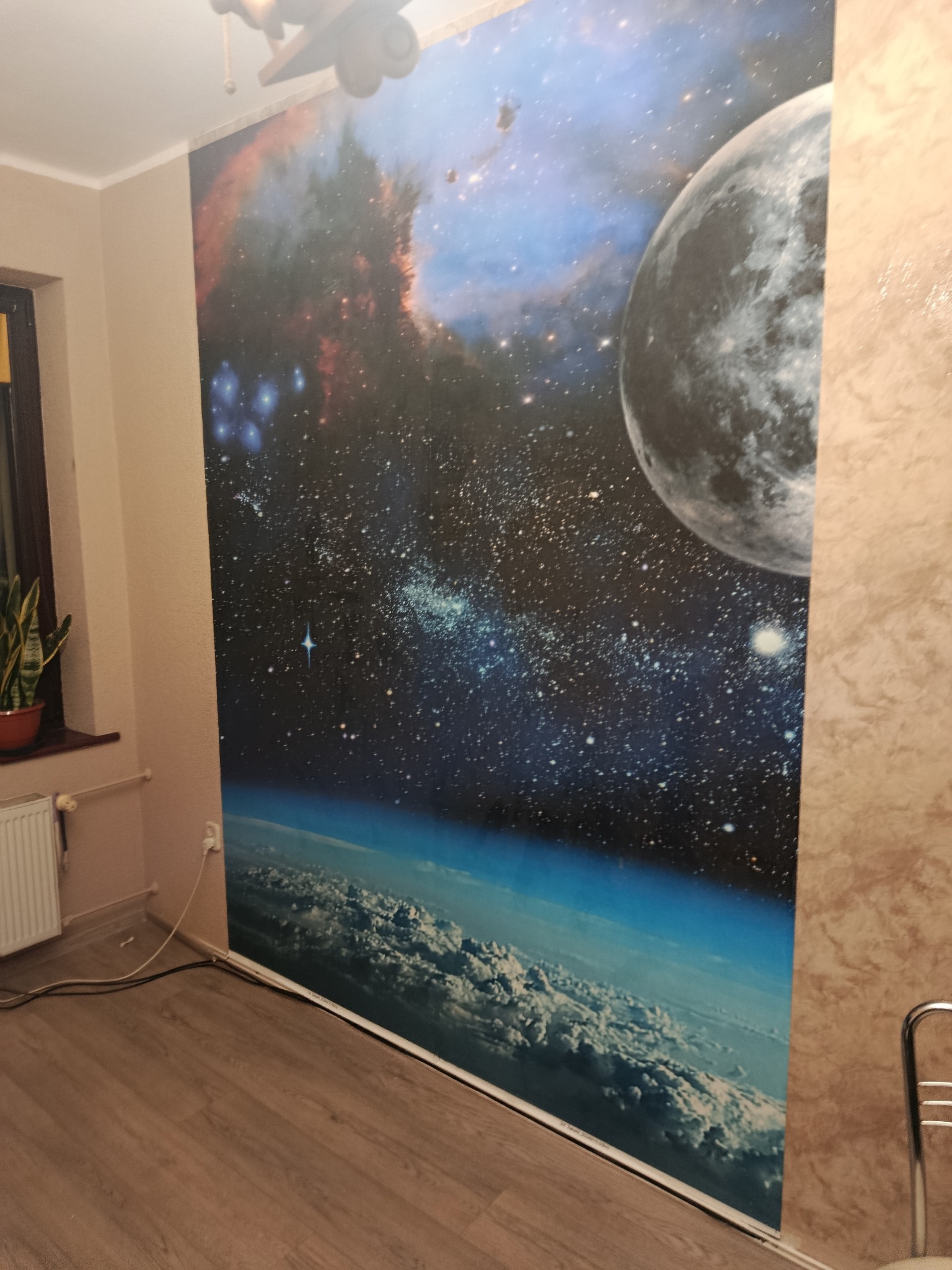 Pokój z fototapetą kosmosu: planeta, gwiazdy, galaktyki. Dekoracja ściany w pokoju z drewnianą podłogą i grzejnikiem. Nietypowy wzór na ścianie.