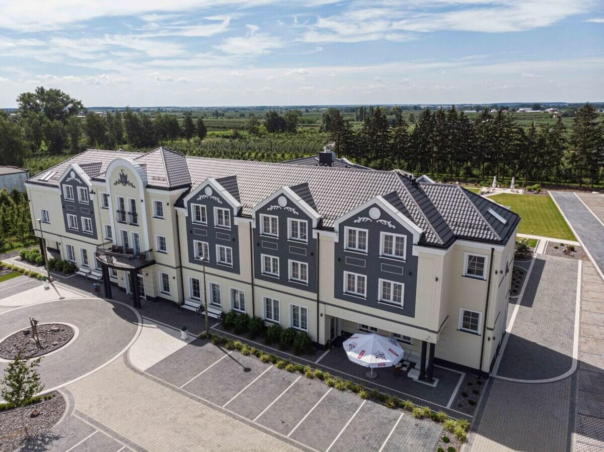 Elewacja budynku z szaro-beżową kolorystyką, widok z lotu ptaka, z przyległym parkingiem i terenem zielonym w tle.