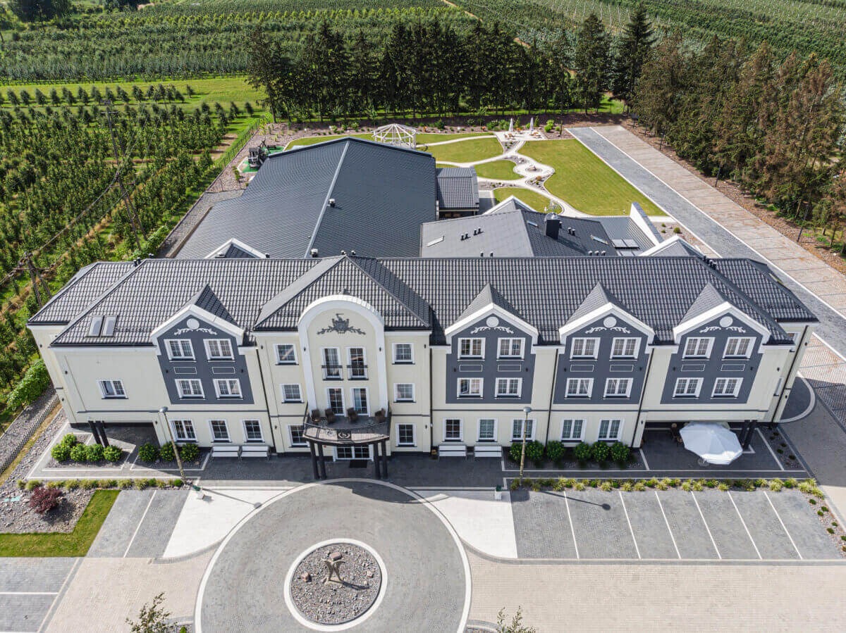 Widok z lotu ptaka na budynek z szarym dachem, jasną elewacją i okrągłym podjazdem z rzeźbą, otoczony zielenią i sadem.