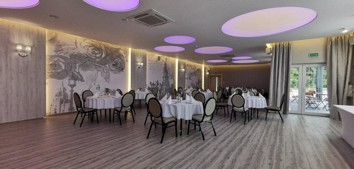 Sala bankietowa z okrągłymi stołami nakrytymi białymi obrusami, szare krzesła, dekoracyjne oświetlenie sufitowe w formie okręgów z fioletową poświatą, na ścianie grafika przedstawiająca motywy...